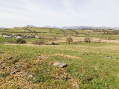 A grassy field with scattered rocks and distant farm buildings surrounded by rolling hills at Orsedd Wen Farmhouse in Betws-Y-Coed