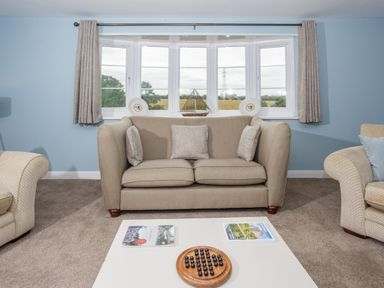 A living room with a beige sofa pillows a white coffee table with books and a wooden game set blue walls and a bay window with curtains at Overbrook in Salwarpe near Droitwich Spa