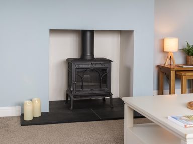 A black wood stove in a fireplace with three candles on the hearth and a wooden table with a lamp and plant nearby at Overbrook in Salwarpe near Droitwich Spa