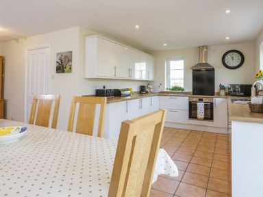 A kitchen with wooden chairs, a table with a polka dot tablecloth, white cabinets, an oven, a clock, and kitchen appliances at Overbrook in Salwarpe near Droitwich Spa