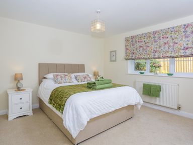 A bedroom with a beige bed white bedding green blankets floral cushions and a window with floral curtains at Overbrook in Salwarpe near Droitwich Spa