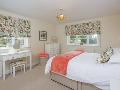 A bedroom with a bed covered in white and orange bedding two windows with floral curtains a white dresser and a white dressing table at Overbrook in Salwarpe near Droitwich Spa