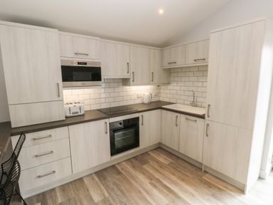 A kitchen with light wood cabinets white tiled backsplash electric stove oven microwave toaster kettle and black chairs at Caban Y Mor Amroth near Saundersfoot