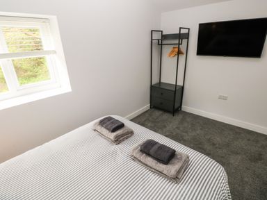 A bedroom with a striped bedspread folded towels a window a clothing rack with hangers and a wall-mounted television at Caban Y Mor in Amroth near Saundersfoot