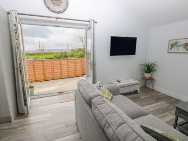 A living room with a gray sofa cushions a wall mounted TV and an open door leading to a patio at Caban Y Mor in Amroth near Saundersfoot