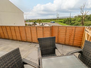 A patio with wicker chairs and a glass-top table enclosed by a wooden fence overlooking a parking area and grassy fields at Caban Y Mor in Amroth near Saundersfoot