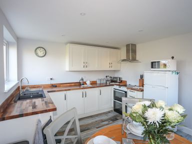 A kitchen with white cabinets wooden countertops a round dining table with flowers and a refrigerator with a microwave on top at Oscars Pad in Brynsiencyn