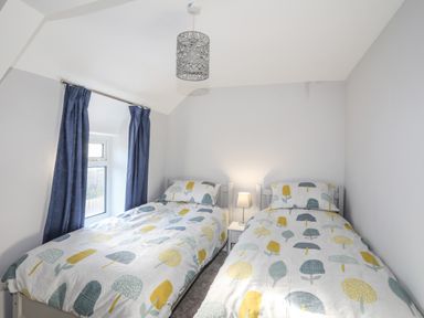A bedroom with two single beds with patterned bedding a bedside table with a lamp and a window with blue curtains at Oscars Pad in Brynsiencyn