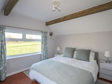 A bedroom with a double bed two bedside tables lamps a window with curtains and wooden ceiling beams at Oscars Pad in Brynsiencyn