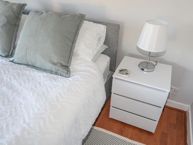A bedroom corner with a bed containing white bedding and green pillows next to a white nightstand with a lamp and a coaster at Oscars Pad in Brynsiencyn