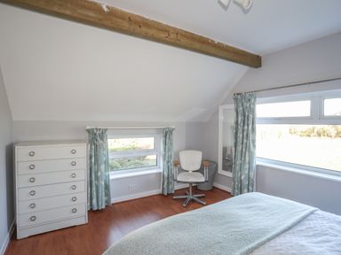 A bedroom with a white chest of drawers a chair near two windows with curtains and a bed with a blanket at Oscars Pad in Brynsiencyn