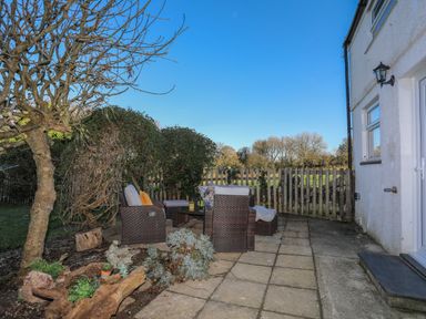 An outdoor patio with wicker chairs and a table next to a house and a wooden fence at Oscars Pad in Brynsiencyn