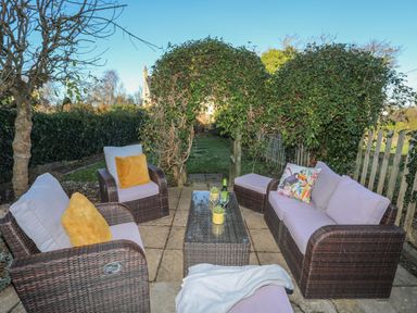 An outdoor patio with wicker chairs and sofa with cushions a glass coffee table with drinks and plants and a leafy archway in the background at Oscars Pad in Brynsiencyn
