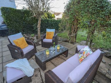 An outdoor patio area with wicker furniture including chairs and a sofa surrounding a glass coffee table with plants and wine glasses at Oscars Pad in Brynsiencyn