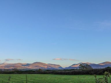 A green field with hedges and trees in front of mountains under a clear blue sky at Oscars Pad in Brynsiencyn