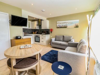 A living room with two gray sofas a wooden dining table a small blue side table a wall mounted television and a kitchenette at Chalet 126a in Cromer