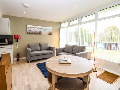 A living room with two gray sofas a round wooden table and large windows with vertical blinds at Chalet 126a in Cromer