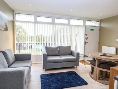 A living room with two gray sofas a blue rug a round wooden dining table and chairs near a door with vertical blinds at Chalet 126a in Cromer