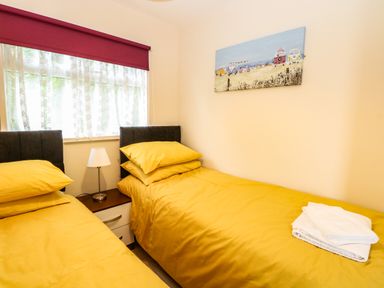 A bedroom with two single beds with yellow bedding a nightstand with a lamp a window with lace curtains and a red blind and a painting on the wall at Chalet 126a in Cromer