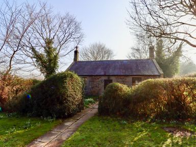A stone cottage with chimney stacks and a slate roof behind a pathway and hedges at Rose Cottage in Akeld near Wooler