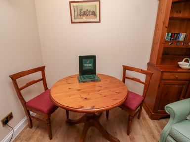 A round wooden table with two wooden chairs with red cushions and a wooden cabinet with books in a room at Rose Cottage in Akeld near Wooler