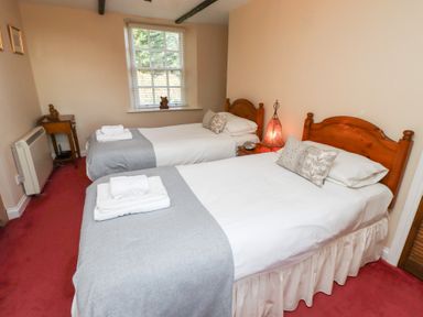 A bedroom with two single beds side by side each with white linens grey throws and pillows a wooden nightstand with a lamp between the beds a window with blinds a small wooden desk and heater on a red carpeted floor at Rose Cottage Akeld near Wooler