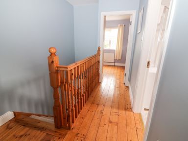 A wooden staircase railing next to a hallway with wooden floors and a doorway leading to a room with a window and curtains at 17 Clover Hill in Killorglin County Kerry