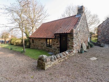 A stone cottage with a red tiled roof and a small stone wall outside in a rural setting at Tile Rouge in Wooler
