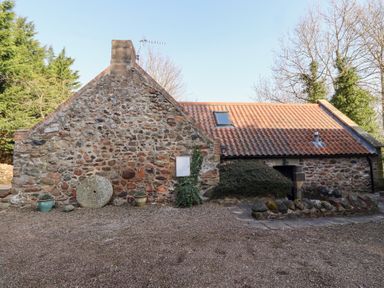 A stone cottage with a tiled roof and gravel yard surrounded by trees at Tile Rouge in Wooler