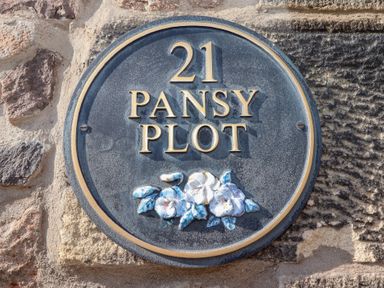 A round plaque with number 21 and Pansy Plot on a stone wall with flower decoration at Pansy Plot Akeld near Wooler