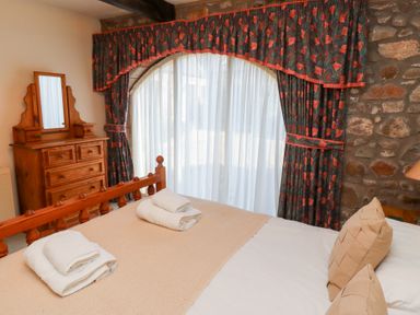 A bedroom with a wooden bed and dresser next to a large window with curtains and a stone wall at Pansy Plot in Akeld near Wooler