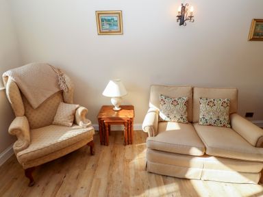 A sitting area with an armchair a loveseat wooden nesting tables with a lamp and framed pictures on the wall at Catkin Cottage Akeld near Wooler