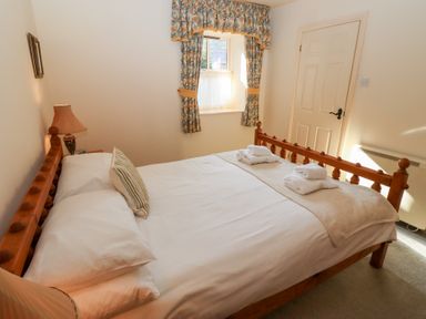 A bedroom with a wooden bed with folded towels on the blanket a window with floral curtains and a closed door at Catkin Cottage in Akeld near Wooler