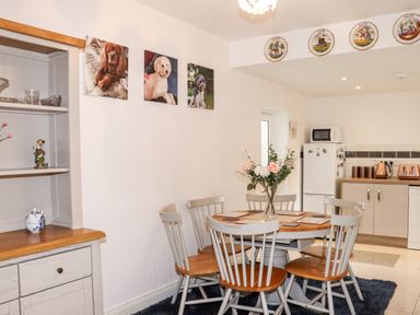 A dining area with a round wooden table and six chairs a flower vase on the table wall art of dogs a kitchen with refrigerator microwave and toaster in the background at Lane Cottage Ballycullane County Wexford
