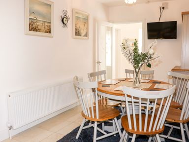 A dining area with a round wooden table and six chairs with light-colored backs a vase with flowers on the table two framed pictures on the wall a wall clock a radiator and a wall-mounted television at Lane Cottage in Ballycullane County Wexford