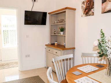A dining area with a round wooden table and white chairs a beige cabinet with shelves and a wall-mounted television in a room at Lane Cottage in Ballycullane County Wexford