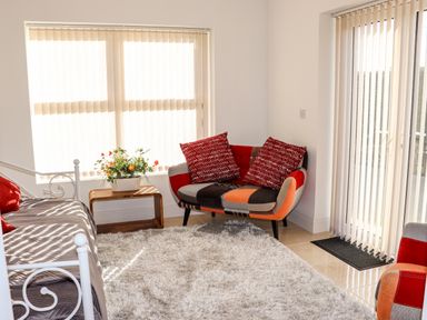 A sitting area with a patchwork loveseat with red pillows a white metal framed daybed a wooden side table with a plant and vertical blinds on windows at Lane Cottage Ballycullane County Wexford