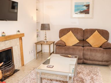 A living room with a brown sofa, yellow cushions, a glass coffee table, a side table with a lamp, a fireplace, and a wall-mounted TV at Lane Cottage in Ballycullane County Wexford