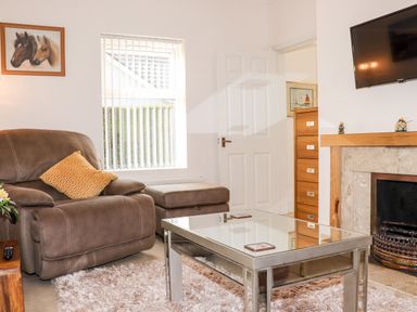A living room with a brown armchair and footstool a glass coffee table a fireplace with mantel and TV mounted above at Lane Cottage in Ballycullane County Wexford
