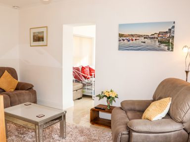 A living room with two brown armchairs a glass coffee table a small wooden side table with flowers a wall painting of boats and a view into another room with a white metal bed and red pillows at Lane Cottage in Ballycullane County Wexford