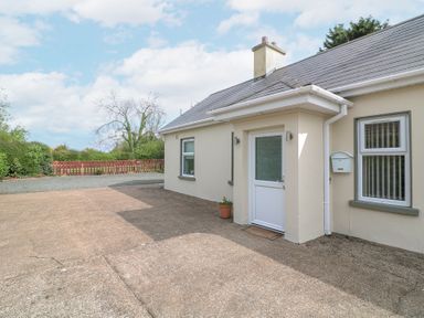 An exterior view of a beige house with a white door a mailbox and windows with vertical blinds surrounded by trees and a wooden fence at Lane Cottage in Ballycullane County Wexford