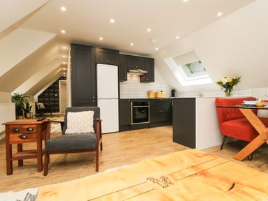 An open-plan kitchen and living area with dark cabinets a white refrigerator a gray chair with a pillow a wooden side table a red upholstered bench and a wooden dining table with a glass top under a slanted ceiling with skylights at Robins Nest at The Shire Inn in Killivose near Camborne