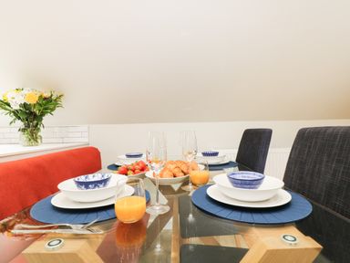 A dining table set with blue placemats white plates blue bowls croissants strawberries orange juice and wine glasses in a dining area with a red bench and dark chairs at Robin's Nest at The Shire Inn in Killivose near Camborne