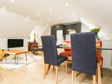 A dining area with navy chairs and a wooden table next to a kitchen with dark cabinets and a living area with a wooden coffee table and a mounted TV at Robins Nest at The Shire Inn in Killivose near Camborne