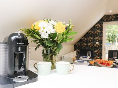 A kitchen counter with a Nespresso machine two cups and a vase of flowers with a dining table set with bowls glasses and cutlery at Robins Nest at The Shire Inn in Killivose near Camborne