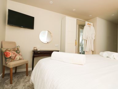 A bedroom with a bed rolled towels a chair with a cushion a wall-mounted television a circular mirror a dark wooden desk and a wardrobe with bathrobes hanging on the door at Robins Nest at The Shire Inn Killivose near Camborne