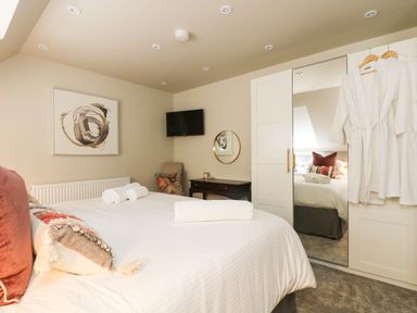 A bedroom with a bed, pillows, rolled towels, wall art, a TV, a chair, a desk, a round mirror, and robes hanging on a wardrobe door at Robins Nest at The Shire Inn in Killivose near Camborne