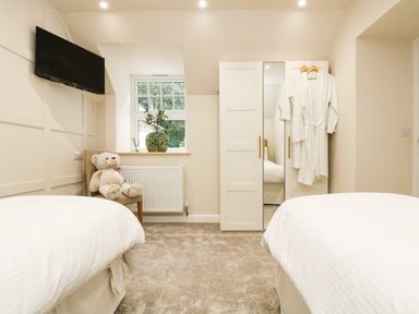 A bedroom with two beds a wardrobe with hanging robes a chair with a teddy bear a window with a vase and a wall-mounted television at Robins Nest at The Shire Inn in Killivose near Camborne