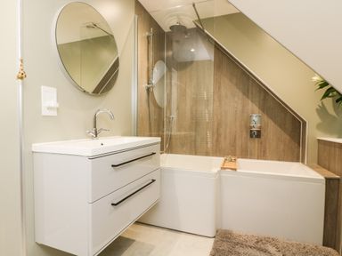 A bathroom with a white sink cabinet a round mirror and a bathtub with a shower under a slanted ceiling at Robins Nest at The Shire Inn in Killivose near Camborne