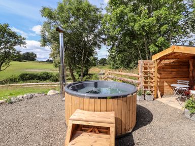 A wooden hot tub with water and steps beside a wooden shelter with a table and chairs outdoors at The Moorhen Retreat in Nantmawr near Trefonen